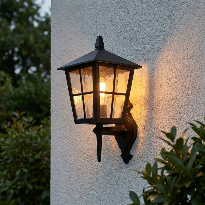 Traditional black lantern wall light with textured glass panes glowing warmly on a white exterior wall.