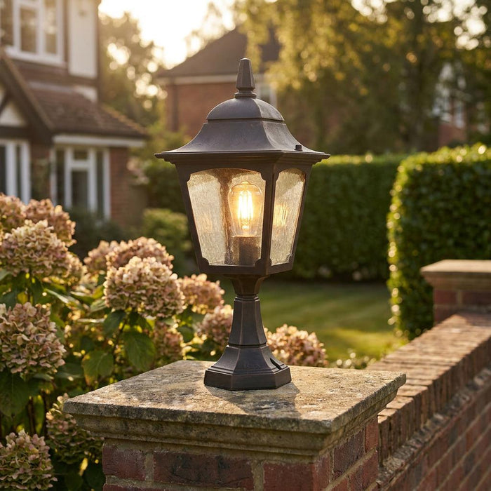 Traditional bronze outdoor pedestal lantern with a warm filament bulb mounted on a stone garden wall pillar.