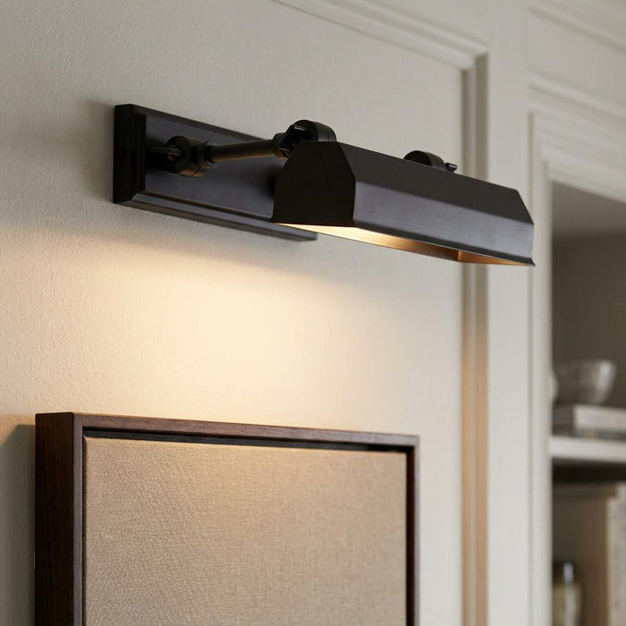 Small black picture light mounted on a wall, casting warm light onto a framed piece of art below.