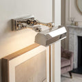 Polished nickel picture light with adjustable twin arms illuminating a framed artwork on a neutral wall.