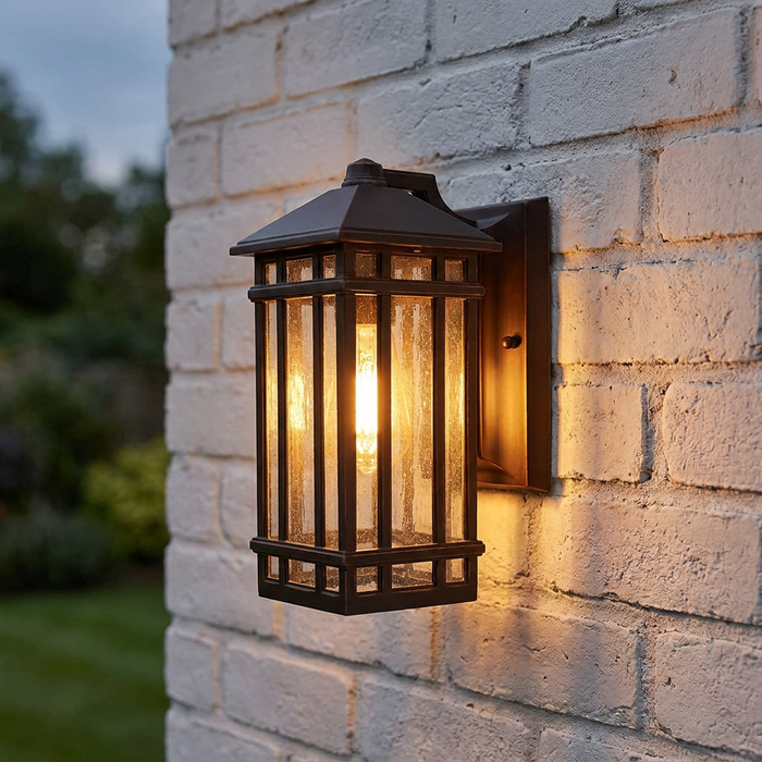A bronze outdoor lantern with seeded glass panes glowing on a white brick wall.