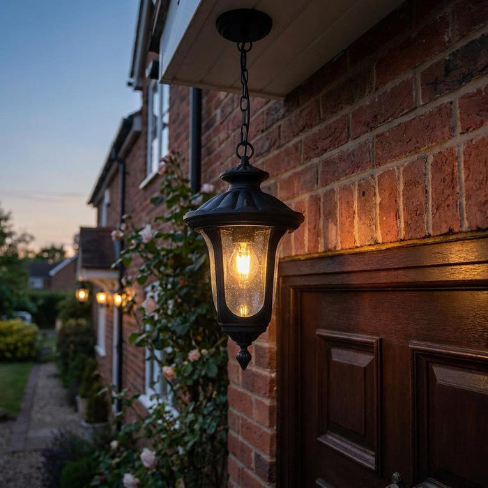 Black hanging chain lantern with a glowing bulb mounted on a brick wall above a wooden door at dusk.