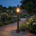 An illuminated Chicago bronze patina outdoor bollard light stands along a brick path in a lush evening garden.
