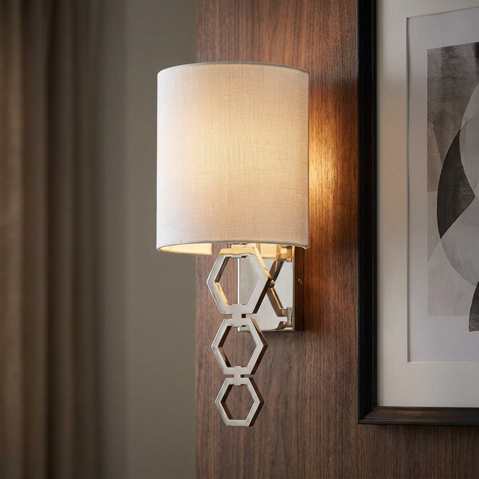 Polished chrome wall light with three hexagonal links and a white drum shade, mounted on a wood panel.