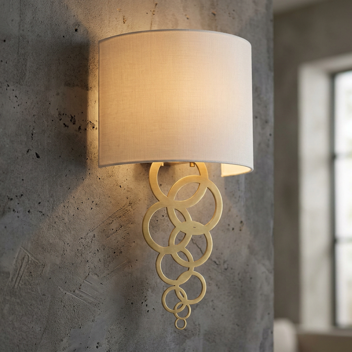 Aged brass wall light with cascading interlocking rings and a white fabric shade on a grey concrete wall.