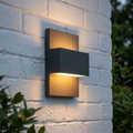 Modern graphite outdoor wall light mounted on a white brick wall, casting a warm upward and downward glow.