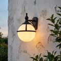 Black globe wall lantern with a white glass shade illuminated on a rustic textured wall.
