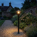 A black outdoor bollard light illuminates a gravel garden path at twilight, surrounded by flowers and greenery.