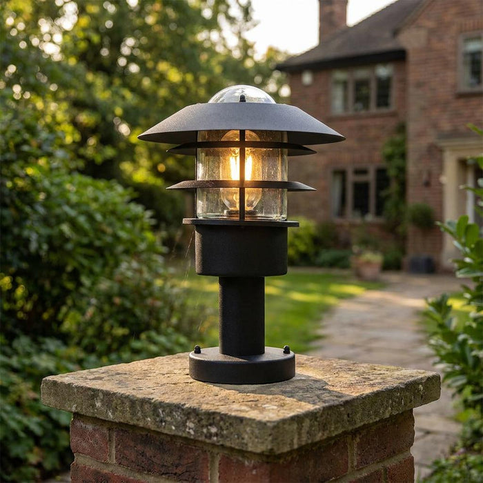 Modern black tiered garden lantern mounted on a brick pillar, illuminated in a garden setting.