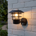Black outdoor wall light with layered metal shades and a glowing bulb, mounted on a white brick wall.
