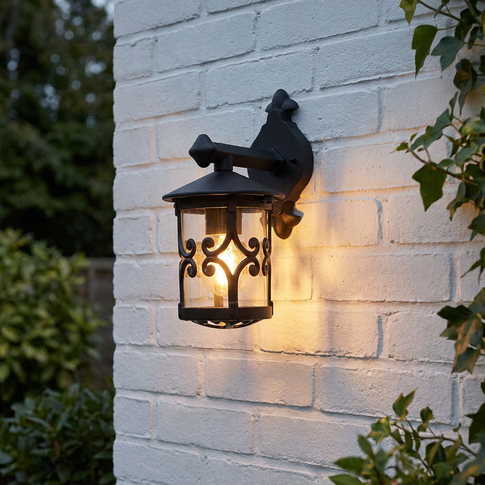 A traditional black outdoor wall lantern with decorative scrollwork glowing on a white brick wall.