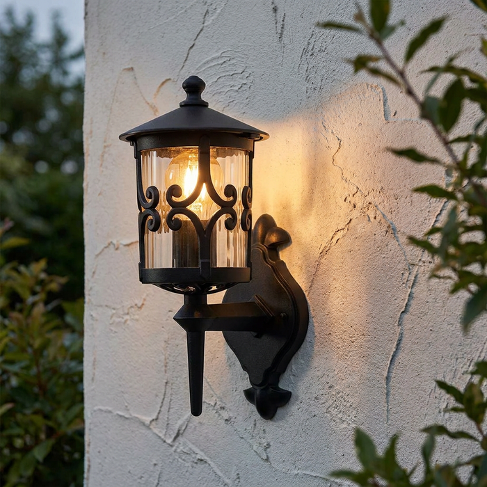 Traditional black outdoor lantern with scrollwork detail, lit on a textured white wall.