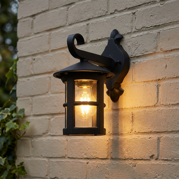 Traditional black outdoor downlighter wall lantern with a warm glowing bulb mounted on a light brick wall.