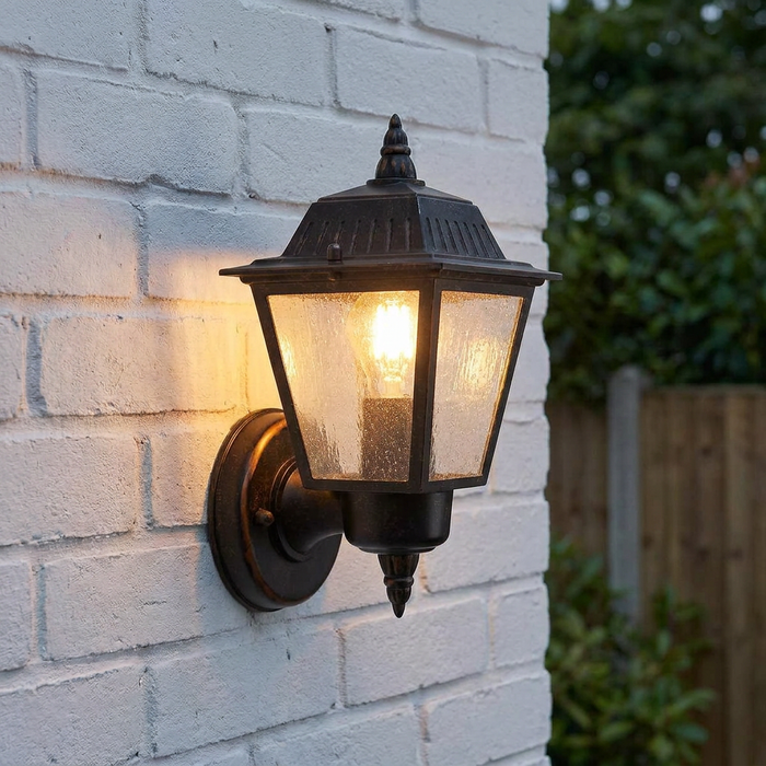 Weathered bronze outdoor wall lantern with seeded glass panels, lit up and mounted on a white brick wall.