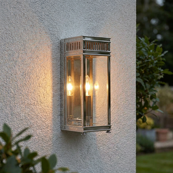 Polished chrome lantern wall light with glowing bulbs mounted on a textured exterior wall.