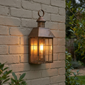 Elstead Hyde Park aged brass lantern wall light with a glowing bulb mounted on a light-colored brick wall.