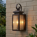 A lit old bronze lantern-style outdoor wall light with seeded glass mounted on a pale brick wall.