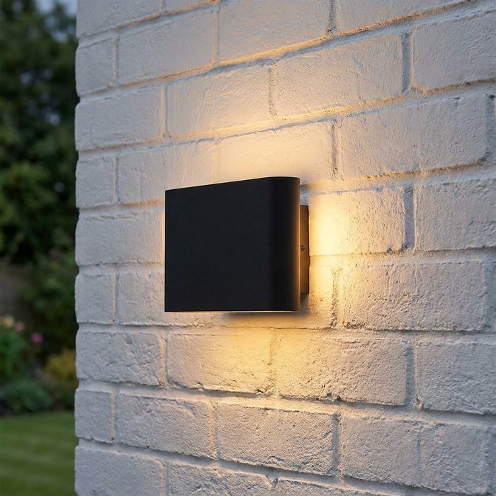Elstead Jens medium black outdoor wall light mounted on a white brick wall, emitting warm light in four directions.