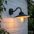 Black curved arm outdoor wall light with a glowing bulb mounted on a white brick wall surrounded by ivy.