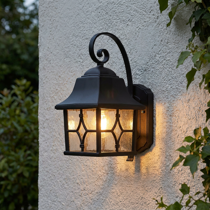 Traditional black outdoor wall lantern with leaded glass panels glowing warmly on a white textured wall with ivy.