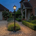 An Elstead Kerry black outdoor lamp post lighting a garden path at dusk near a brick house.