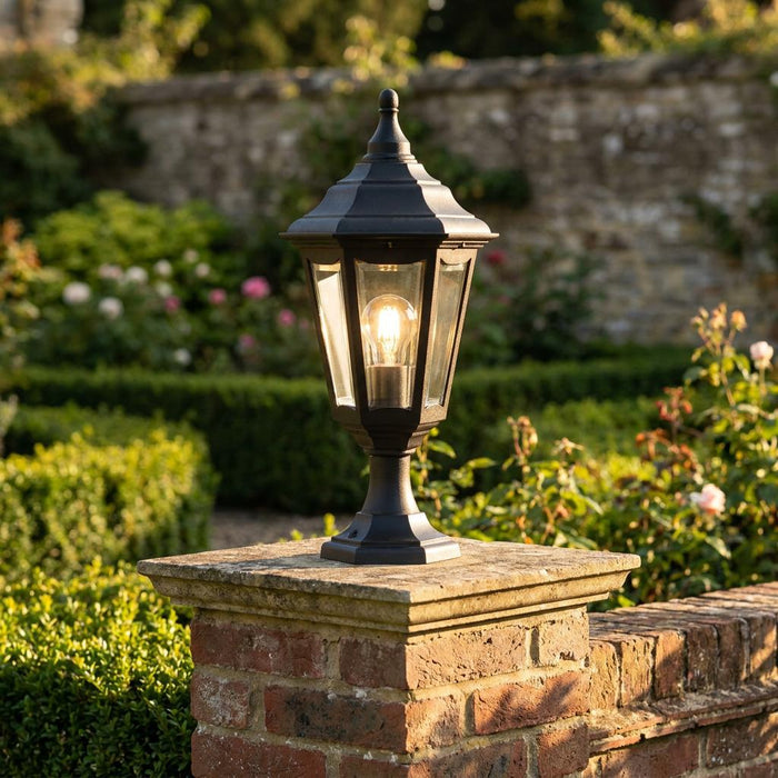 Lit black traditional hexagonal lantern mounted on a brick garden wall pillar in a lush garden setting.