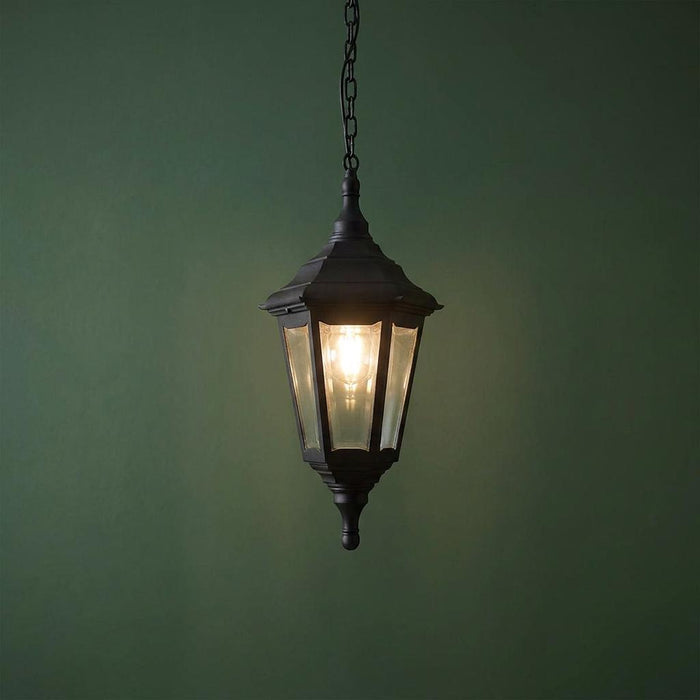 Traditional black outdoor lantern pendant light with clear glass panes hanging against a dark green wall.
