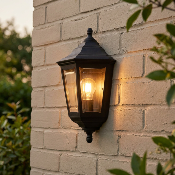A black traditional outdoor lantern wall light with a glowing warm bulb on a white brick wall.