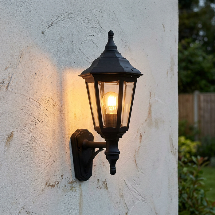 Traditional black outdoor wall lantern with a warm glowing bulb, mounted on a white textured exterior wall.