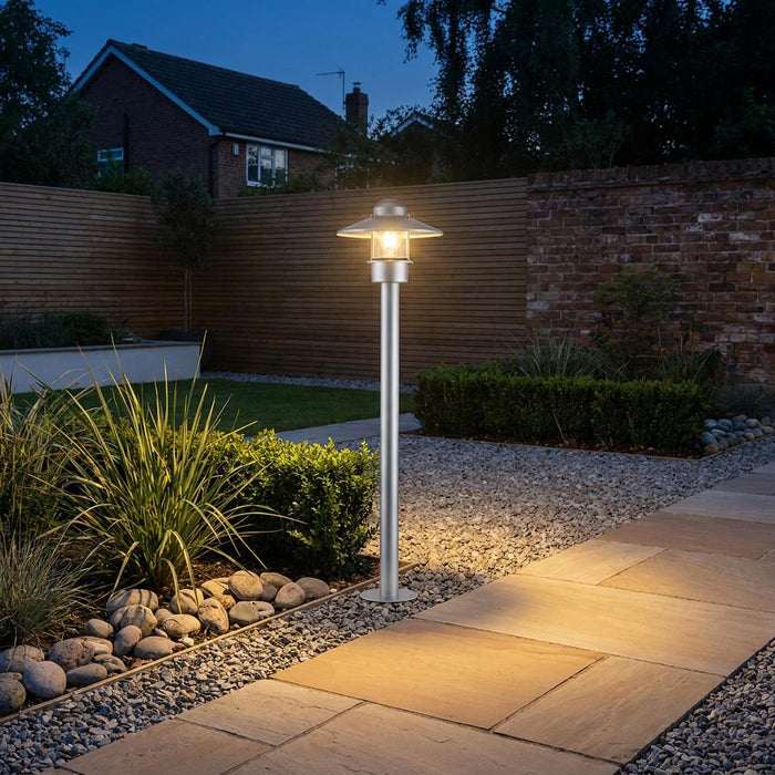 Silver outdoor bollard light illuminating a stone garden path at dusk near a wooden fence and shrubs.
