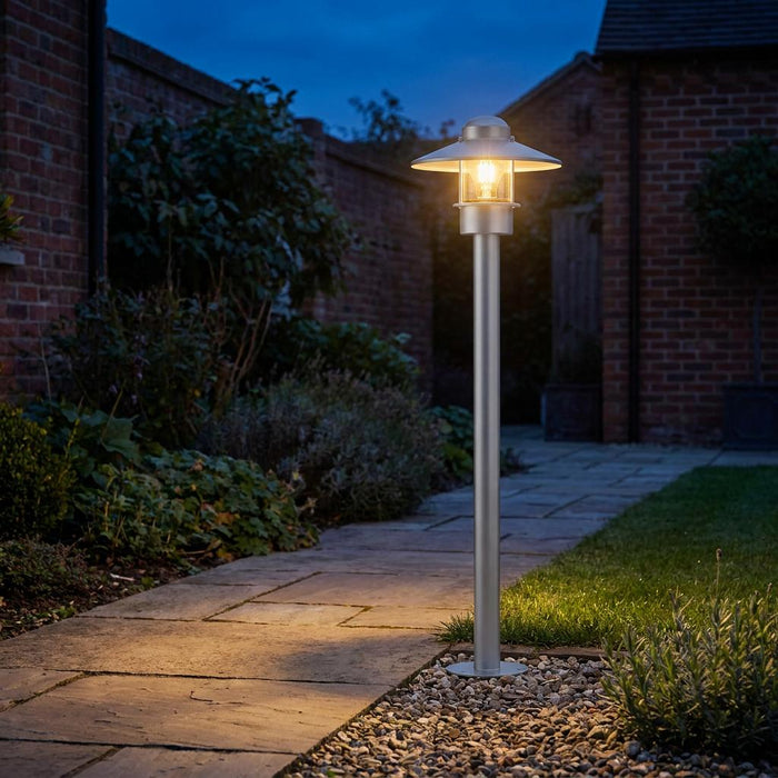 Silver outdoor bollard light illuminating a stone garden path at dusk with a warm glowing bulb.