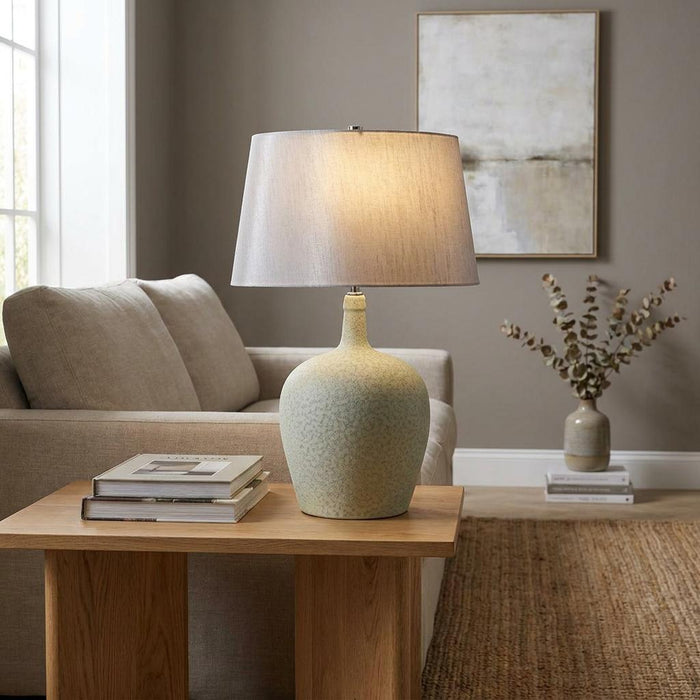 Sage green ceramic table lamp with a grey fabric shade on a wooden side table in a modern living room.