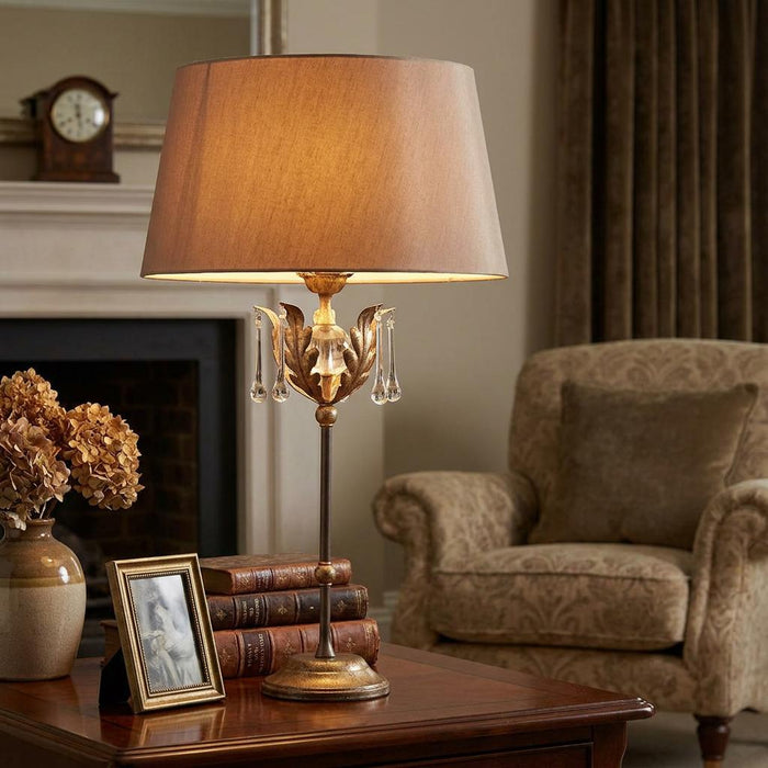 Bronze table lamp with ornate leaf details and crystal droplets glowing on a wooden table in a living room.
