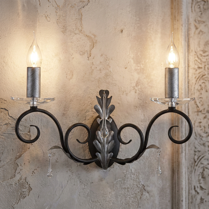Two-light wall sconce with black and silver scrollwork, crystal drops, and candle bulbs on a textured plaster wall.