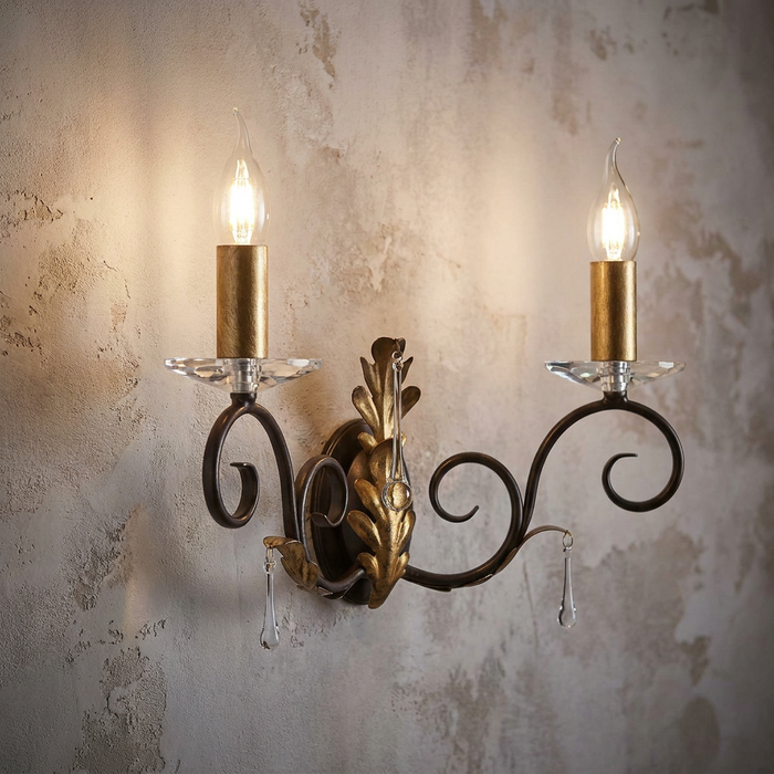 Scrolled bronze Amarilli two-light wall sconce with gold leaf and crystal droplets lit on a textured wall.