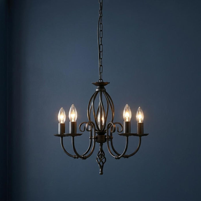 Five-light black wrought iron chandelier with lit candle bulbs and scrollwork against a dark blue wall.