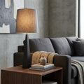 Black geometric table lamp with a grey textured shade on a wooden side table in a modern living room.