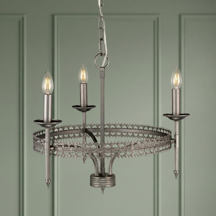 Silver 3-light crown chandelier with intricate metal scrollwork, hanging in front of a green paneled wall.