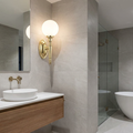 Polished brass wall light with a white globe shade mounted on a grey tiled bathroom wall near a mirror.