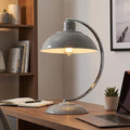 Grey industrial desk lamp with a curved chrome arm illuminating a wooden desk in a modern office setting.