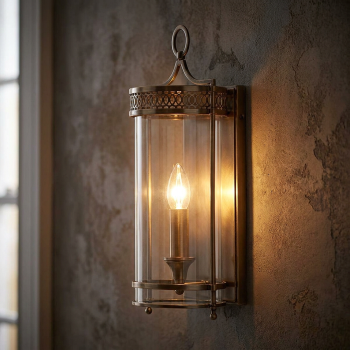 Dark bronze wall light with a clear cylindrical shade and lit candle bulb mounted on a rustic textured wall.