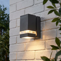 An illuminated Jannik Graphite Outdoor Wall Light with a textured rectangular body and clear glass windows on a white brick wall.
