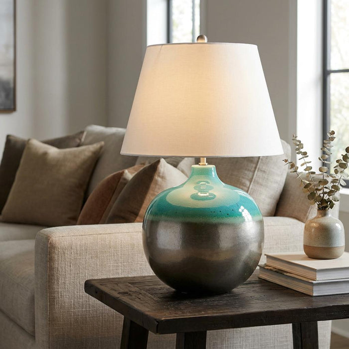 Large ceramic table lamp with a teal and metallic bronze base and white shade on a side table in a living room.
