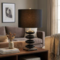 Black ceramic pebble-base table lamp with a dark fabric drum shade on a wooden side table in a modern living room.