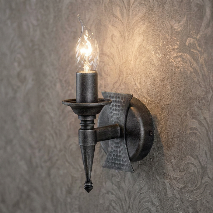 Black candle-style wall light with hammered metal detail and a glowing flame bulb on damask wallpaper.