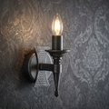 Medieval-style black and silver wall sconce with a lit candle bulb against a dark damask wallpaper.