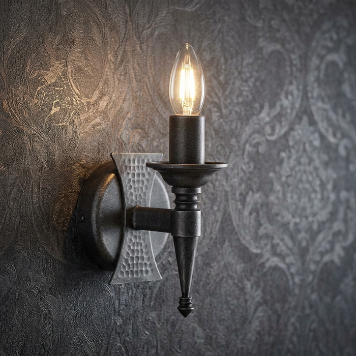 Medieval-style black and silver wall sconce with a lit candle bulb against a dark damask wallpaper.