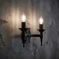 Medieval-style two-light black wall sconce with lit candle bulbs on a distressed grey concrete wall.