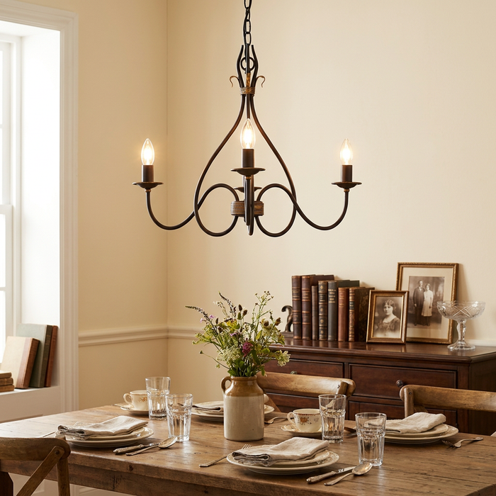 Three-light rust and gold chandelier with elegant scrolls illuminating a set wooden dining table in a cozy room.