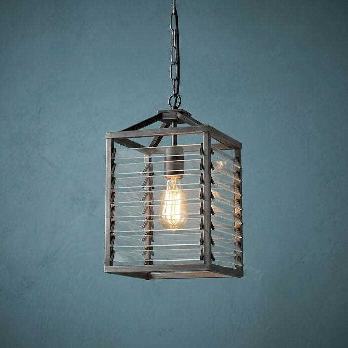 Gunmetal lantern-style pendant light with horizontal glass slats and a glowing bulb against a textured blue wall.
